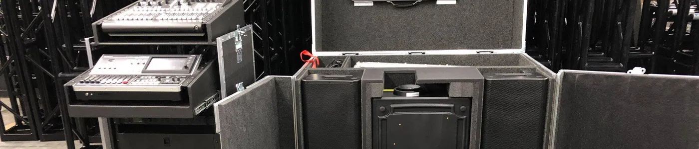 Black open road case in an AV warehouse showing two rack-mounted control surfaces—a video switcher and media player—on the left, and compartments housing a flat-panel monitor, telescoping mounting arms, and accessories on the right, with stacks of black steel truss segments visible behind