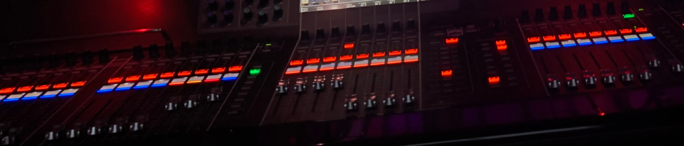 Consoles_LPG Close-up of a professional LED lighting console showing multiple motorized faders with illuminated red and blue status buttons, encased in a glossy black panel under low ambient light