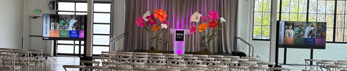Two on-stage confidence monitors on black-draped pedestals—one displaying the event hashtag ‘#RWON’ with colorful geometric branding, the other showing a four-up presenter view above a countdown timer—positioned at the front edge of a conference stage alongside a microphone stand and floor monitors