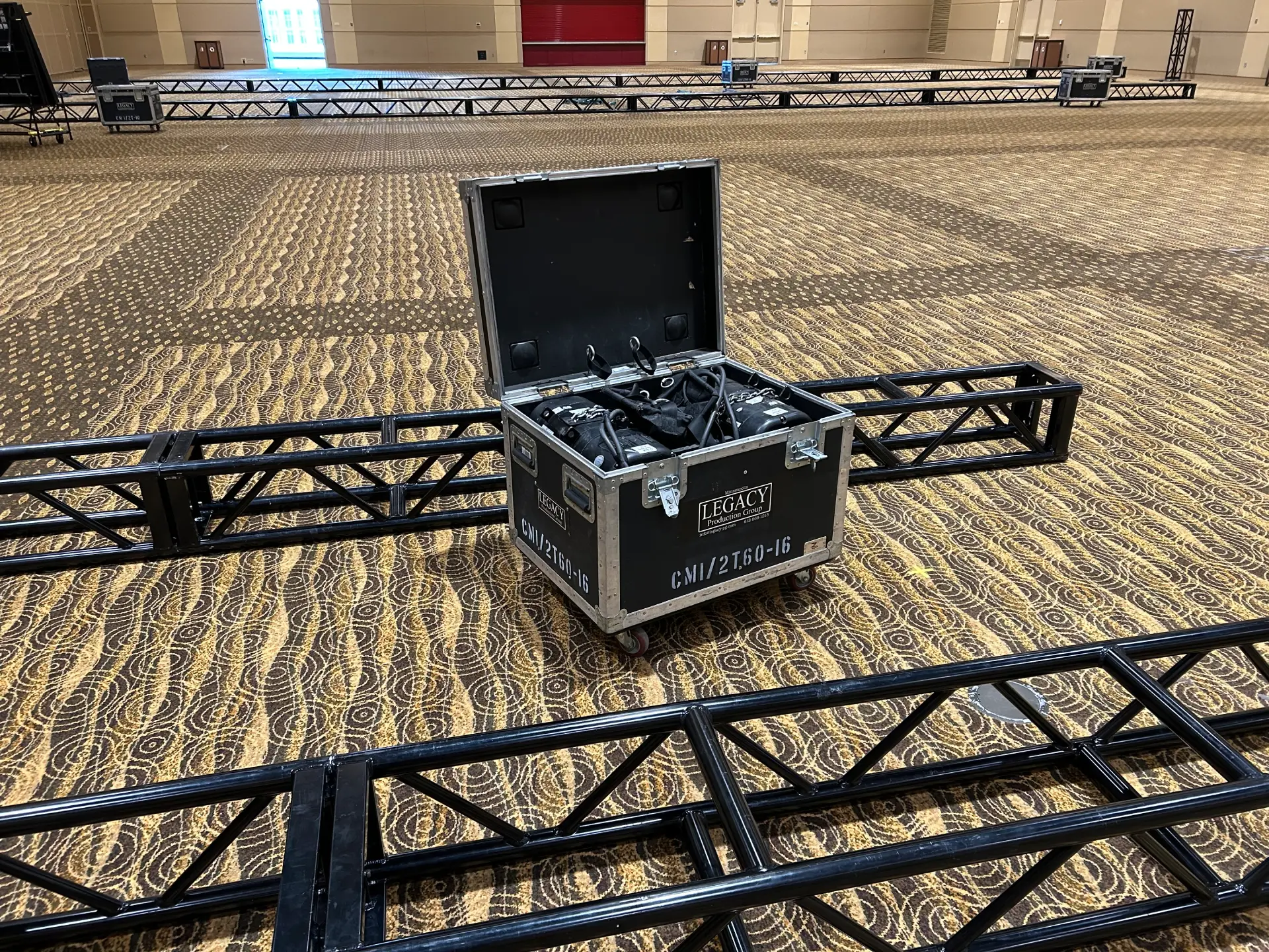Large warehouse space with dozens of black steel truss segments arranged in parallel rows on wheeled stands across a concrete floor, ready for event rigging installations