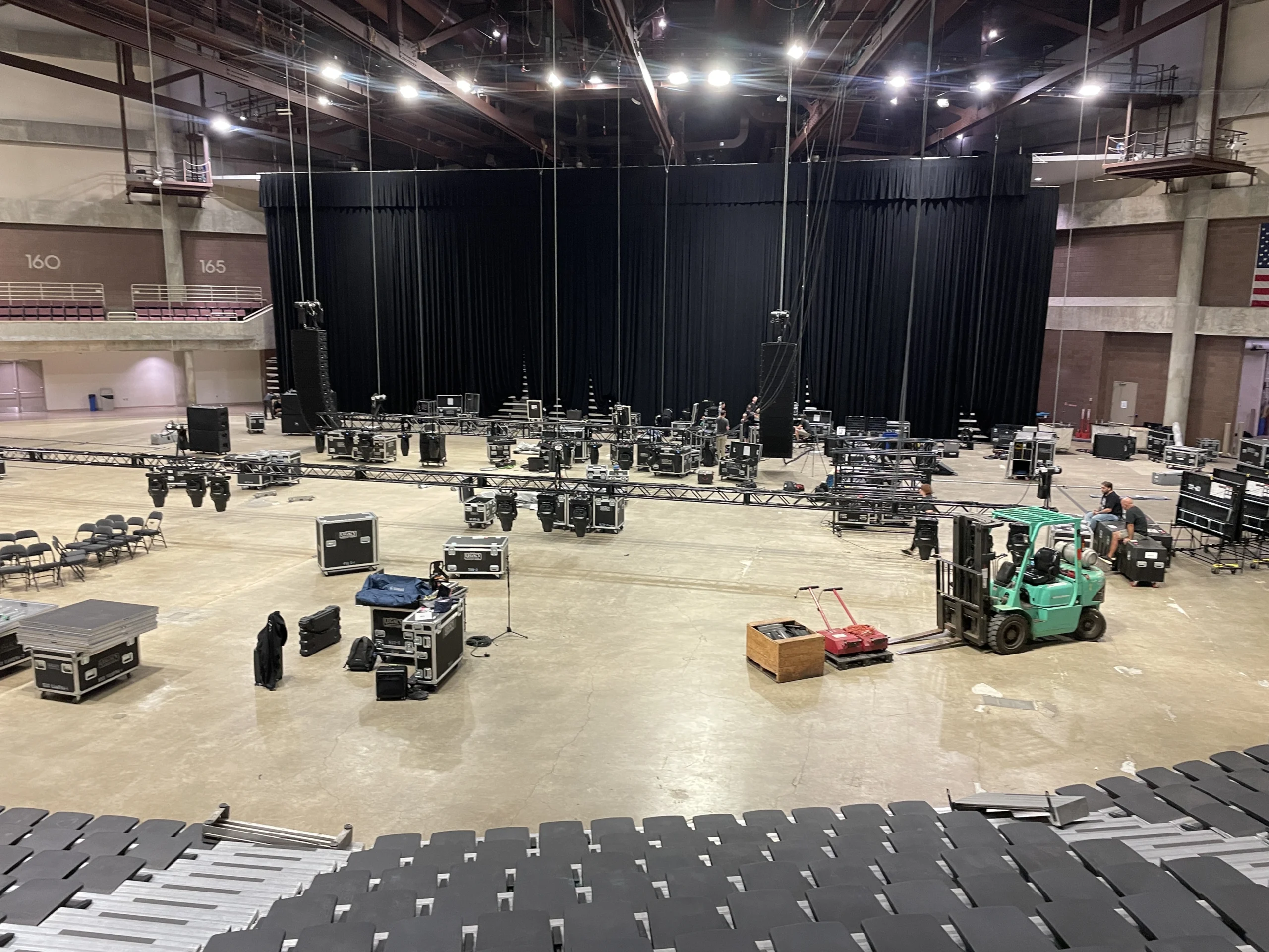 Overhead view of an arena floor during event load-in showing a green forklift beside black stage truss sections, flight cases, lighting fixtures, and folded riser panels on the concrete slab; a large black drape backdrop spans the far end beneath numbered seating sections