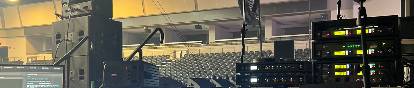 Microphones-and-IEMs Backstage production station view inside an arena, featuring a video preview monitor at left, a floor monitor speaker beside it, and a rack of wireless microphone receivers with illuminated front panels at right, all overlooking rows of empty seats and overhead lighting trusses ready for live event production