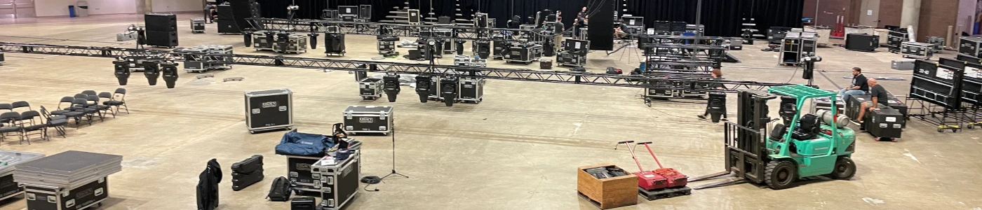 Arena event load-in scene on a concrete floor with a green forklift moving Legacy Production Group flight cases, scattered black truss sections and lighting fixtures, and technicians organizing AV equipment beneath a large black drape backdrop and overhead rigging