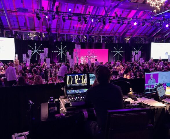 Wide-angle view of a hotel ballroom banquet setup with dozens of round tables dressed in white linens and black chairs under dynamic gobo patterns and blue-and-purple uplighting, all facing a front stage framed by flown truss lighting and multiple colored LED video screens displaying branded graphics, ready for a gala dinner presentation