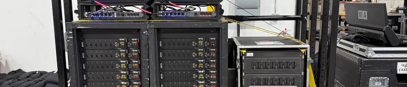 Legacy Production Group mobile power distribution rack showing black rack-mounted distro panels with rows of 120 V outputs labeled by circuit and blue IEC stage-power connectors, topped by audio interface units, all mounted on a steel frame with swivel casters in an AV warehouse setting