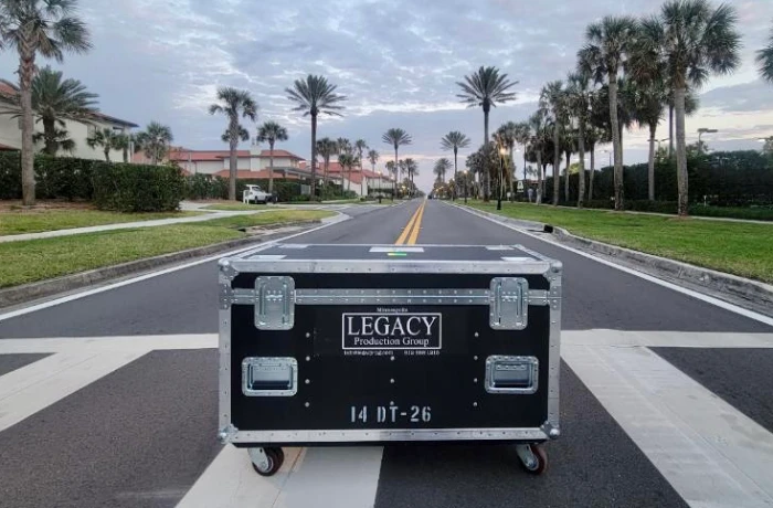 Black wheeled road case labeled ‘LEGACY Production Group 14 DT-26’ positioned in the middle of a palm-lined suburban street at dusk, showcasing branded AV transport solutions against a backdrop of low-rise residential buildings and an overcast sky