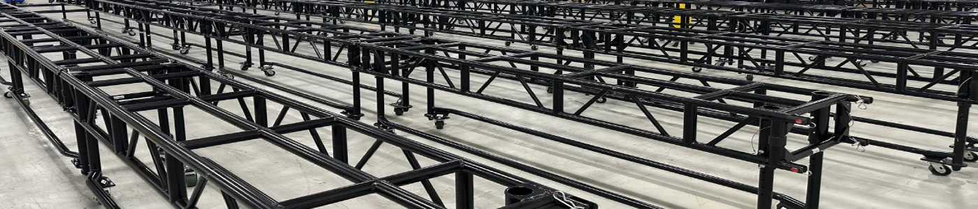 Large warehouse space with dozens of black steel truss segments arranged in parallel rows on wheeled stands across a concrete floor, ready for event rigging installations