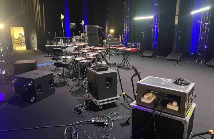 Indoor performance stage setup on a black floor featuring a drum kit with cymbals, a red keyboard on stand, stage monitors, microphone stands, and audio flight cases with cable looms, all beneath trusses bathed in blue uplighting and linear LED fixtures