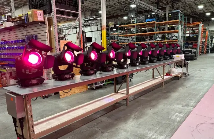 Warehouse staging area with a long metal workbench holding a row of black moving-head stage lights emitting magenta wash; industrial shelving stocked with AV flight cases and truss sections fills the background