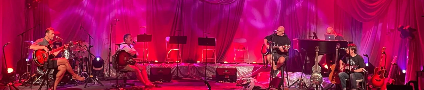 Live acoustic band seated across a draped stage bathed in red uplighting, featuring guitarists, a keyboardist, and microphone stands in front of flowing red curtains and subtle gobo light patterns above
