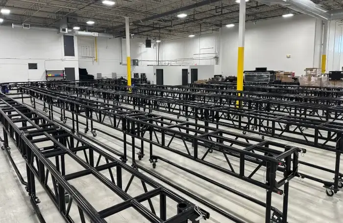 rigging equipment preparing for event Large warehouse space with dozens of black steel truss segments arranged in parallel rows on wheeled stands across a concrete floor, ready for event rigging installations