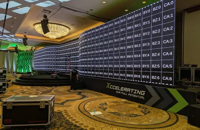 Curved indoor stage in a hotel ballroom featuring a large multi-panel LED video wall displaying a test-pattern grid with alphanumeric panel IDs, a branded stage skirt reading ‘Xcelerating Our Full Potential,’ and a technician with a ladder working beneath a crystal chandelier over patterned carpet